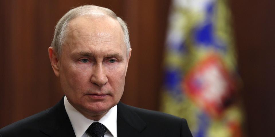 Russian President Vladimir Putin standing in front of a Russian flag while wearing a black suit with a patterned tie.