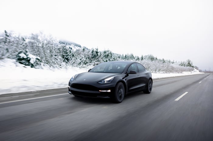 A black Tesla car driving on an open road with a snowy landscape in the background.