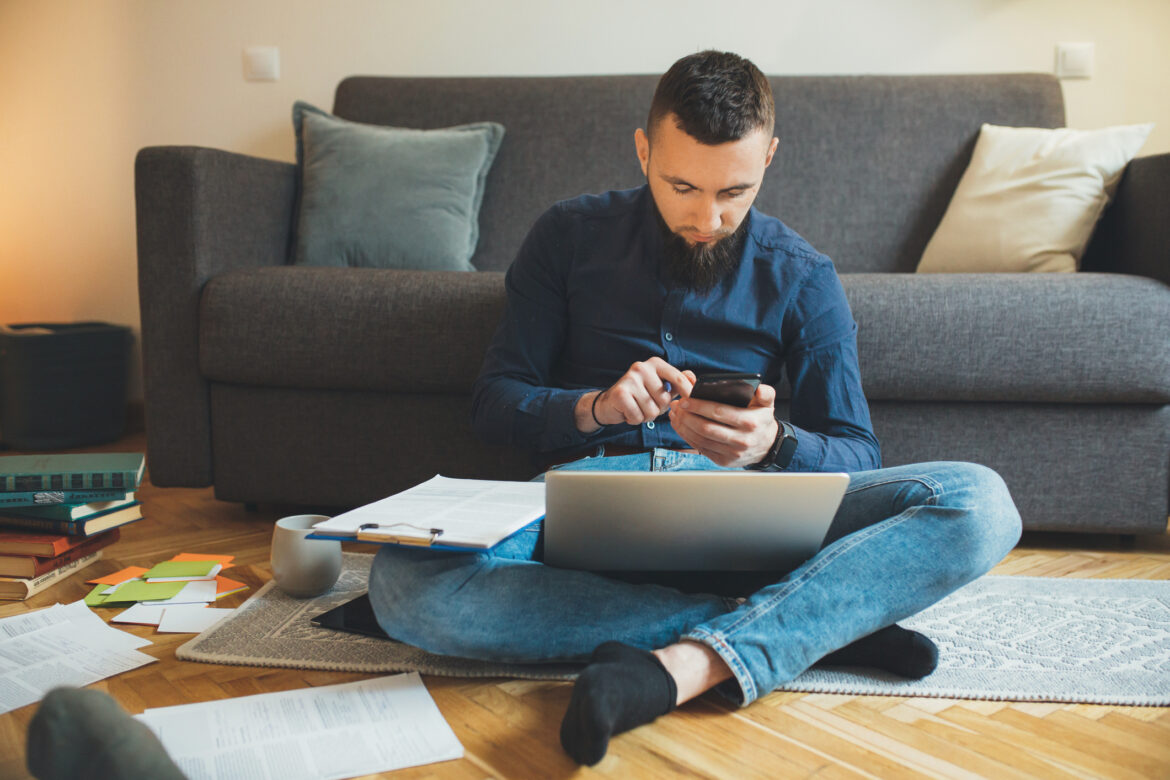 man-sitting-on-floor-holding-phone-with-laptop-and-clipboard-in-lap_gettyimages-1208697647.jpg
