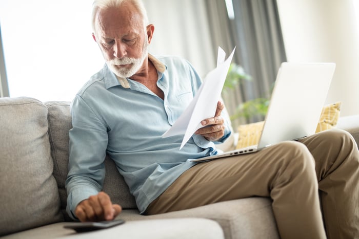 A person sitting on a couch with a laptop holding papers and using a calculator.