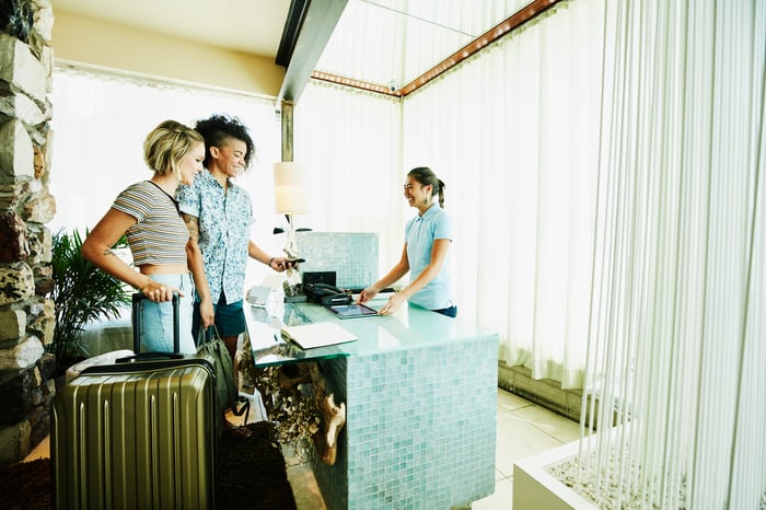 Two people with luggage stand at a hotel checkout desk. 