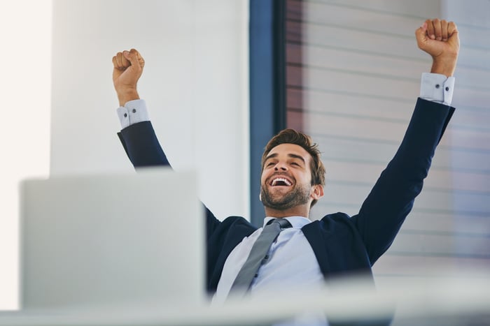 Smiling man in suit at laptop raising arms in celebration