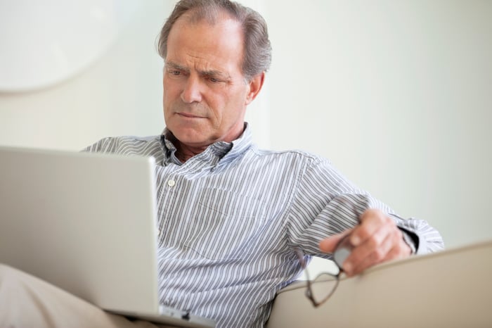 A person seated on a couch holding their glasses while critically reading material on an open laptop.