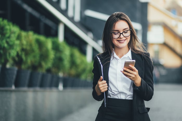 A smiling investor walks down a city street and looks at her phone. 