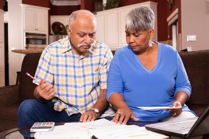 A couple seated on a couch who are closely examining bills and financial paperwork on a table in front of them.