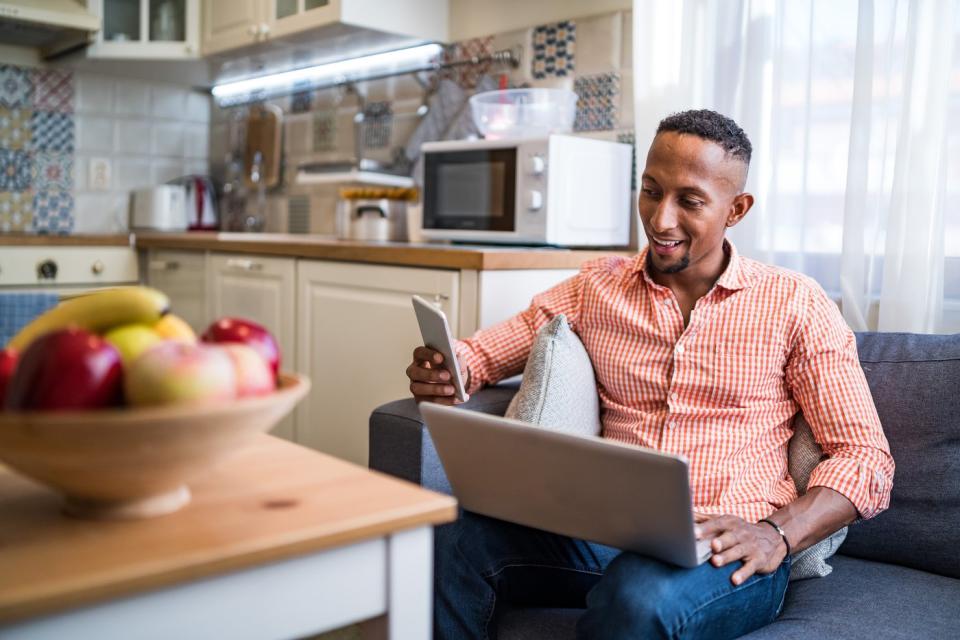 A person seated in their home who's looking at a smartphone in their right hand, with an open laptop on their lap.