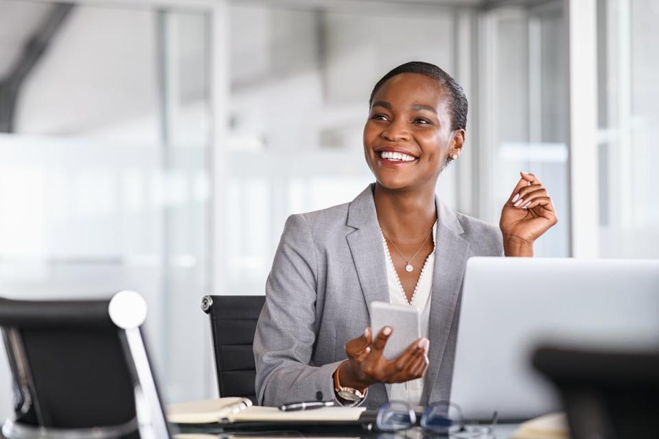 A person smiles while holding a phone and sitting in front of a laptop. 