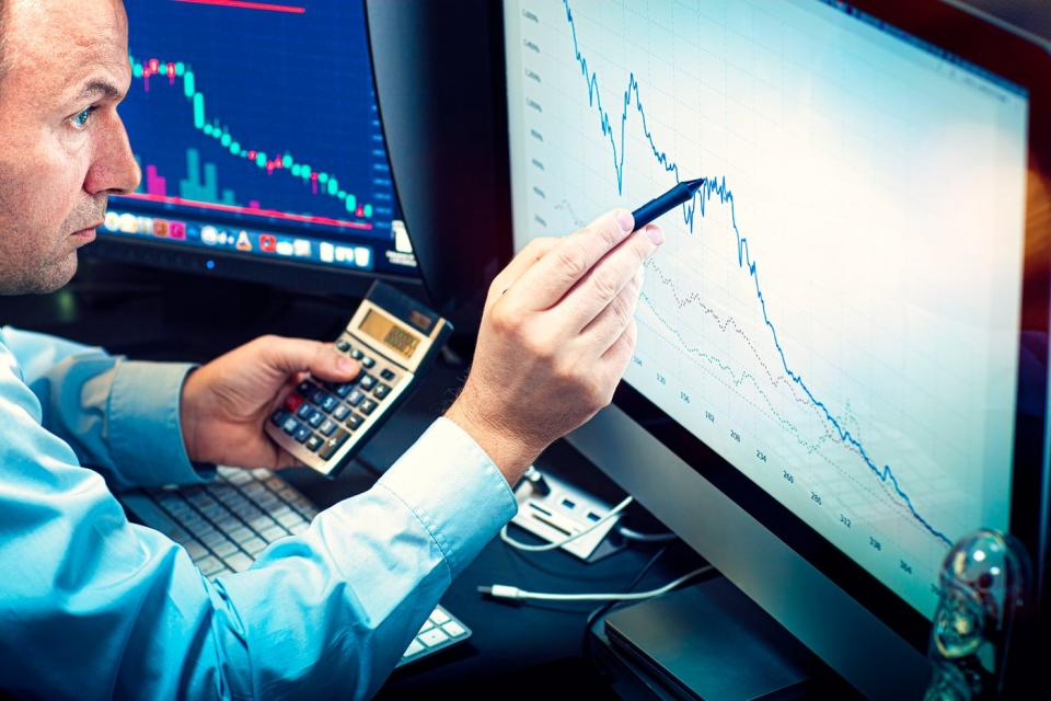 A money manager using a calculator and pen to analyze a plunging stock chart displayed on a computer screen.