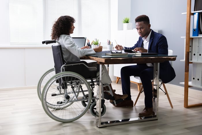 Two people sitting across a table from each other holding writing utensils and looking at documents.