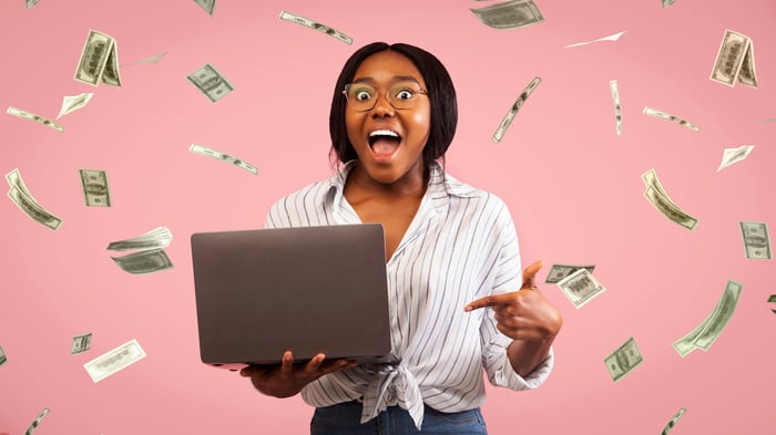 Smiling person holding laptop, with rain of cash falling on them.