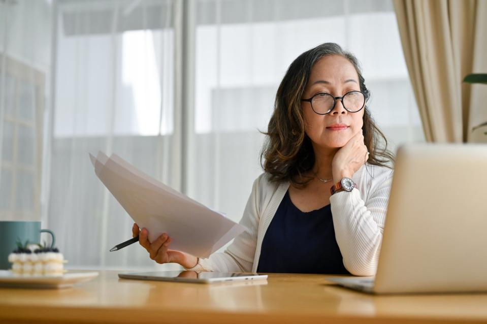 A businessperson looking at an open laptop while holding paperwork in their right hand.